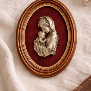 Peut inclure: Sculpture en relief encadrée ovale représentant une mère et son enfant. Les figures argentées se détachent sur un fond rouge foncé, dans un cadre de couleur bronze. Une œuvre d'art religieuse ou sentimentale.