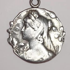 Puede incluir: Colgante plateado con un perfil clásico femenino dentro de un marco circular, adornado con una corona de laurel. El colgante tiene un lazo en la parte superior para colgar. El metal presenta un aspecto ligeramente envejecido.