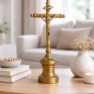 Puede incluir: Un crucifijo dorado con una representación detallada de Jesús en la cruz. La cruz está montada sobre una base decorativa. El crucifijo se exhibe sobre una mesa de madera, con un jarrón y un cuenco con objetos en el fondo.