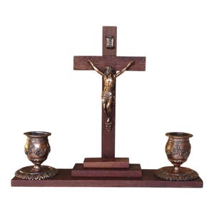 Peut inclure: Un crucifix en bois foncé avec une figure en bronze de Jésus-Christ. Deux bougeoirs en bronze avec des motifs floraux sont de chaque côté. Le crucifix et les bougeoirs sont sur une base en bois brun foncé.