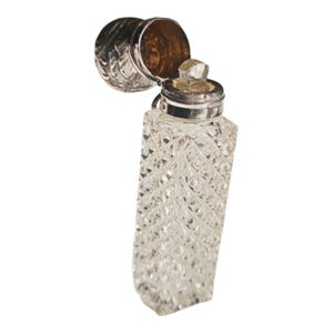 Peut inclure: Un flacon de parfum rectangulaire transparent avec un motif taillé en diamant. Le flacon a un bouchon en métal argenté et un couvercle à charnière. Le couvercle est ouvert, révélant l'intérieur. Le flacon est probablement vintage ou antique.