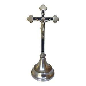 Puede incluir: Un crucifijo plateado con una detallada figura de Jesucristo. La cruz tiene extremos redondeados decorativos y una incrustación negra. Se apoya sobre una base plateada ensanchada, para una exhibición religiosa.