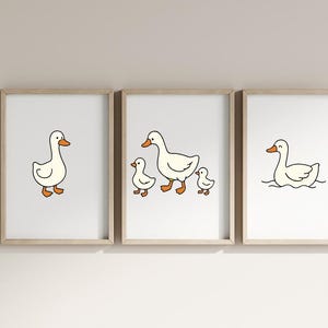 K&ouml;nnte beinhalten: Drei gerahmte Drucke mit Cartoon-Enten. Ein Druck zeigt eine einzelne Ente, ein anderer eine Entenmutter mit zwei K&uuml;ken, und der dritte zeigt eine schwimmende Ente. Die Enten sind wei&szlig; mit orangefarbenen Schn&auml;beln und F&uuml;&szlig;en, schwarz umrandet.