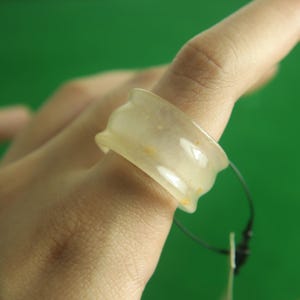 May include: A translucent, light yellow ring with a wavy design. The ring is worn on a finger and appears to be made of a smooth, possibly stone-like material. The background is a solid green.