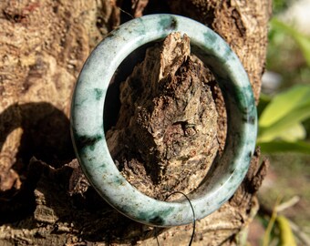 May include: A jade bangle bracelet with a marbled appearance, featuring shades of green and black. The bracelet is circular and smooth, showcasing natural variations in color. The bangle is displayed on a piece of textured wood, highlighting its organic aesthetic.