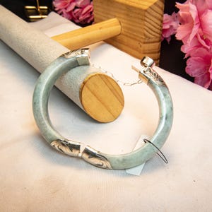 May include: A light green jade bangle bracelet with silver accents. The bracelet features ornate silver detailing at the ends and a silver chain. The jade has a marbled appearance. The bracelet is displayed on a white surface.