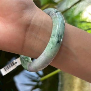Op de afbeelding: Een jade armband in groen- en grijstinten, gedragen om een pols. Het gladde, gepolijste oppervlak van de armband reflecteert licht en toont de natuurlijke kleurvariaties. Het ontwerp van de armband is eenvoudig en elegant.
