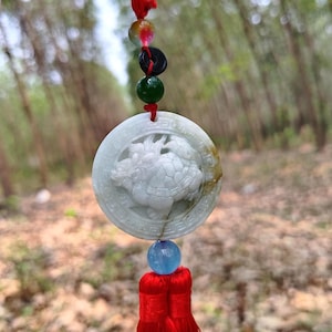 May include: A round, light green jade pendant with a carved dragon and turtle design. The pendant is suspended by a red string with colorful beads and a red tassel. The pendant is approximately 2 inches in diameter.
