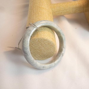 Könnte beinhalten: Ein hellgrünes Jade-Armreif-Armband. Das runde Armband hat eine polierte Oberfläche mit Marmoreffekt, die Farbvariationen aufweist. Eine silberne Kette und ein Anhänger sind befestigt. Ein klassisches Schmuckstück.