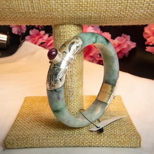 May include: A jade bangle bracelet with silver accents and a red gemstone. The bracelet has a light green and white color with silver filigree detailing. The clasp is gold-colored. A small silver chain hangs from the silver accent.