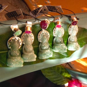 May include: A collection of jade Buddha pendant necklaces. Each pendant features a seated Buddha figure with a silver halo and a gemstone accent. The gemstones are either green or red. The pendants are displayed on a green leaf.