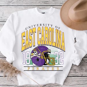 May include: White sweatshirt featuring the text "UNIVERSITY OF EAST CAROLINA" in yellow, with a purple and gold football helmet graphic. The number "1932" is below the text, and the word "PIRATES" is at the bottom.