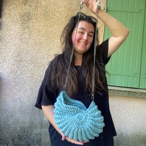 Peut inclure: Un sac à main en crochet bleu clair fait à la main avec une bandoulière en chaîne argentée. Le sac a un design texturé unique et est tenu par une personne. C'est un accessoire élégant.