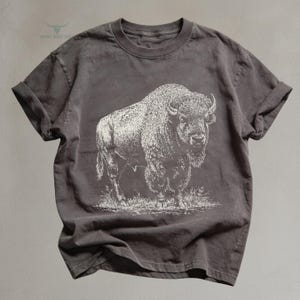 May include: A charcoal gray t-shirt featuring a detailed white illustration of a bison. The bison is standing in a grassy area. The shirt has rolled-up sleeves and a crew neck.