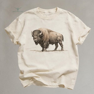 May include: Cream-colored t-shirt featuring a detailed illustration of a bison. The bison is depicted in shades of brown and tan, standing on a grassy area. The shirt has short sleeves and a crew neck. The text "QUIET WEST TEES" is printed on the upper left.