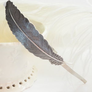 Puede incluir: Una diadema de plumas plateada con un diseño detallado. La diadema está hecha de metal y tiene una base fina y de alambre.