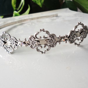Op de afbeelding: Een zilverkleurige haarband met een sierlijk, symmetrisch ontwerp. Het centrale gedeelte heeft een bloemmotief met wervelende details en kleine kristallen. De haarband is geschikt voor speciale gelegenheden of bruidskleding.