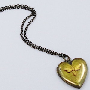 Puede incluir: Un collar con un dije en forma de corazón dorado con un diseño de abeja. La cadena es de metal oscuro.