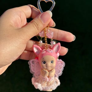 May include: A pink keychain featuring a small doll with bunny ears, a pink outfit, and delicate wings. The doll has its hands clasped in front of its chest. The keychain includes a gold heart-shaped clasp and decorative pink beads.