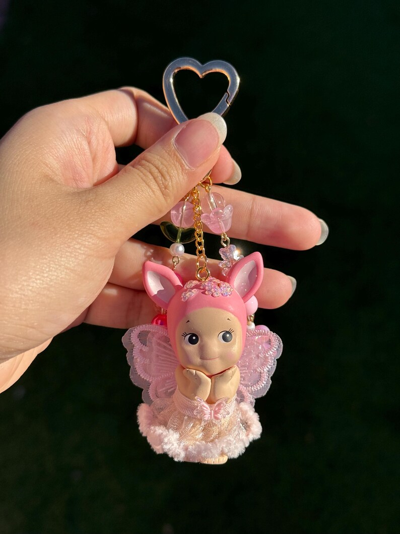 May include: A pink keychain featuring a small doll with bunny ears, a pink outfit, and delicate wings. The doll has its hands clasped in front of its chest. The keychain includes a gold heart-shaped clasp and decorative pink beads.