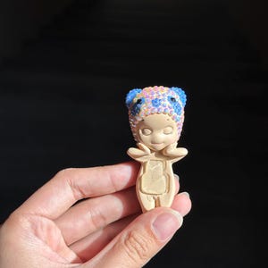 May include: A small, beige, standing figurine with closed eyes and hands touching its cheeks. The figurine wears a headpiece covered in iridescent, multi-colored rhinestones, including blue, pink, and yellow gems, with two rounded ears on top.