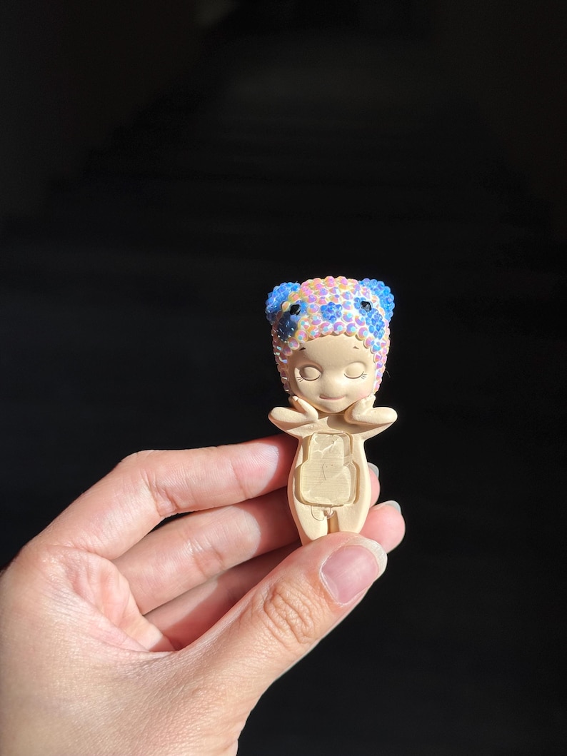 May include: A small, beige, standing figurine with closed eyes and hands touching its cheeks. The figurine wears a headpiece covered in iridescent, multi-colored rhinestones, including blue, pink, and yellow gems, with two rounded ears on top.