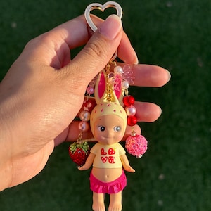 May include: A keychain featuring a small figurine with a yellow bunny-eared hat, a yellow shirt with the red text LOVE, and a pink skirt. The figure is accented with dangling beaded strands, including red and pink spheres and a strawberry charm.