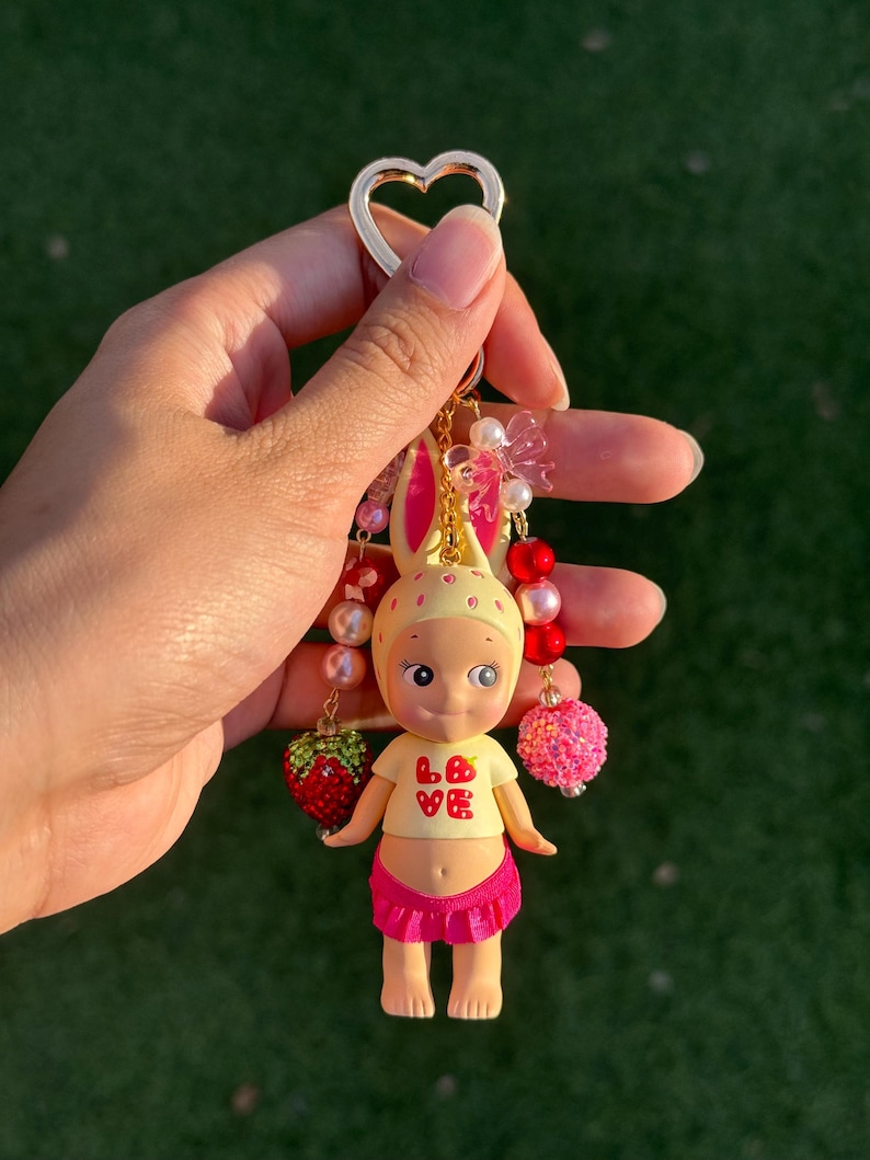 May include: A keychain featuring a small figurine with a yellow bunny-eared hat, a yellow shirt with the red text LOVE, and a pink skirt. The figure is accented with dangling beaded strands, including red and pink spheres and a strawberry charm.