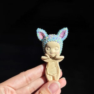 May include: A small, beige figurine with closed eyes and hands touching its cheeks. It wears a cap with pointed ears, entirely covered in iridescent, light-reflecting sequins. The figurine has a smooth, matte finish on its body and face.
