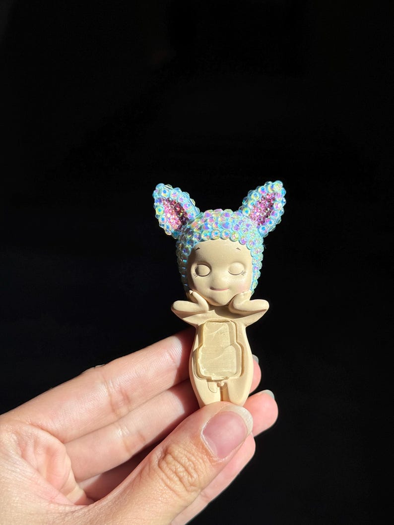 May include: A small, beige figurine with closed eyes and hands touching its cheeks. It wears a cap with pointed ears, entirely covered in iridescent, light-reflecting sequins. The figurine has a smooth, matte finish on its body and face.
