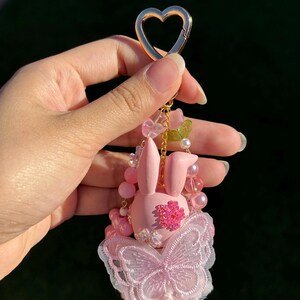 May include: A pink bunny keychain with a gold heart-shaped clasp. The bunny has a flower on its chest and a butterfly-shaped lace design. The keychain is adorned with pink beads and small flowers.