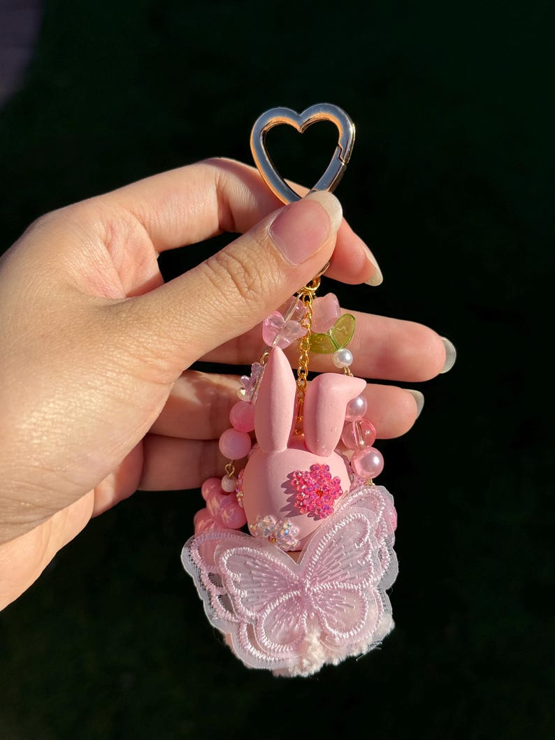 May include: A pink bunny keychain with a gold heart-shaped clasp. The bunny has a flower on its chest and a butterfly-shaped lace design. The keychain is adorned with pink beads and small flowers.