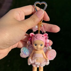 Puede incluir: Un llavero rosa con una peque&ntilde;a mu&ntilde;eca vestida de hada cerdito. La mu&ntilde;eca tiene un gorro de cerdo rosa, alas y un vestido rosa y dorado. El llavero tiene un cierre dorado en forma de coraz&oacute;n y est&aacute; adornado con cuentas y abalorios.