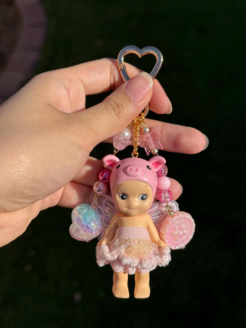 Puede incluir: Un llavero rosa con una peque&ntilde;a mu&ntilde;eca vestida de hada cerdito. La mu&ntilde;eca tiene un gorro de cerdo rosa, alas y un vestido rosa y dorado. El llavero tiene un cierre dorado en forma de coraz&oacute;n y est&aacute; adornado con cuentas y abalorios.