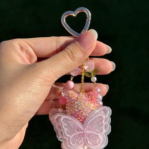 May include: A pink and iridescent keychain with a heart-shaped clasp. The keychain features a butterfly design with a pink sequined bodice, a pink embroidered butterfly, and pink beaded accents. A charming accessory.