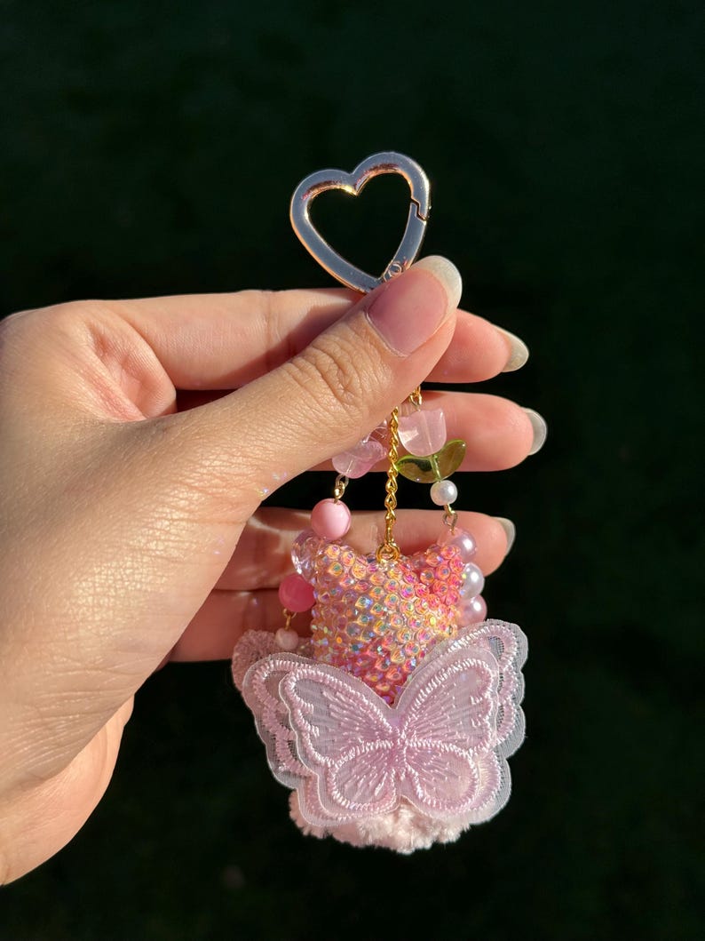 May include: A pink and iridescent keychain with a heart-shaped clasp. The keychain features a butterfly design with a pink sequined bodice, a pink embroidered butterfly, and pink beaded accents. A charming accessory.