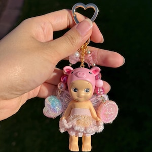 Puede incluir: Un llavero caprichoso con una mu&ntilde;eca de hada cerdito. La mu&ntilde;eca tiene un gorro de cerdo rosa, un tut&uacute; rosa y blanco, y alas iridiscentes. El llavero incluye un cierre en forma de coraz&oacute;n y cuentas decorativas.