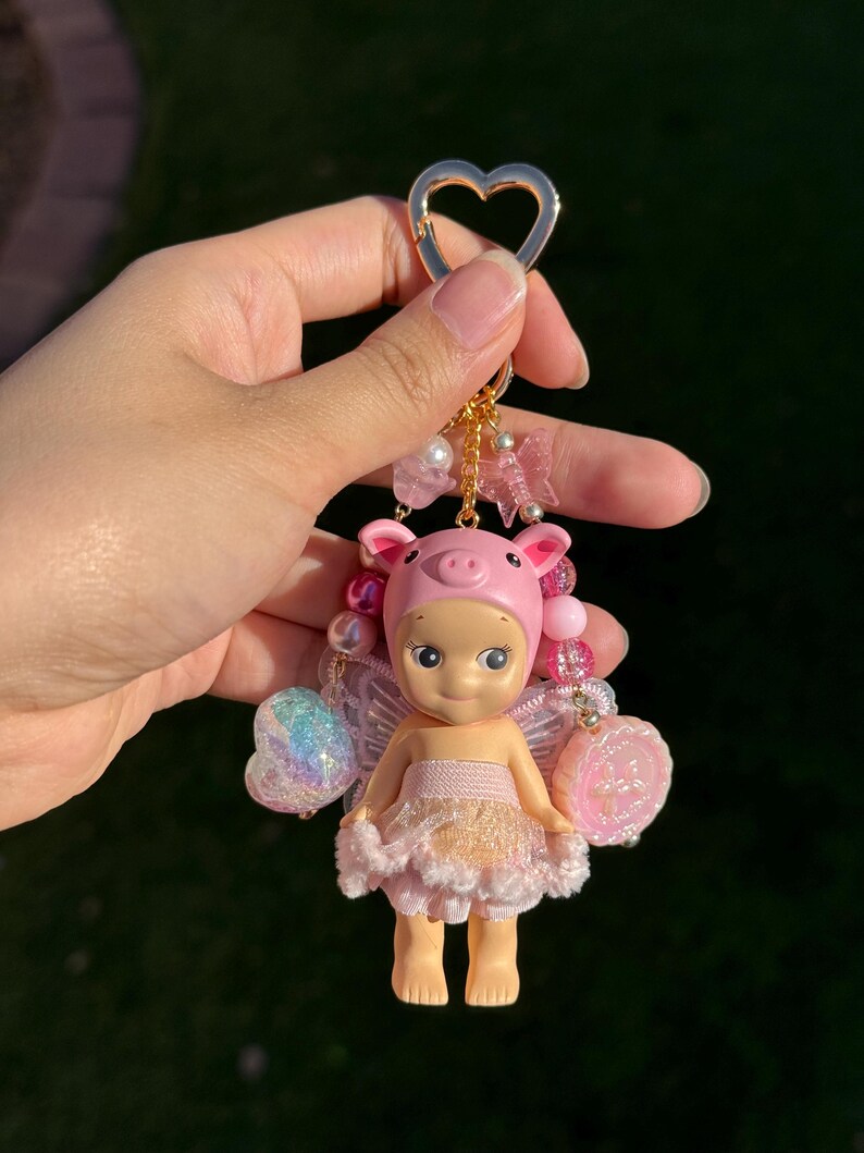 Puede incluir: Un llavero caprichoso con una mu&ntilde;eca de hada cerdito. La mu&ntilde;eca tiene un gorro de cerdo rosa, un tut&uacute; rosa y blanco, y alas iridiscentes. El llavero incluye un cierre en forma de coraz&oacute;n y cuentas decorativas.