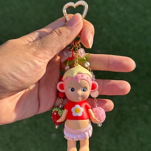 May include: A keychain featuring a small figurine with blonde hair, a pink strawberry-patterned headpiece, and a red shirt with a white flower graphic. The figure wears a light pink ruffled skirt and is accented with dangling beads, a strawberry charm, and a heart-shaped gold clasp.