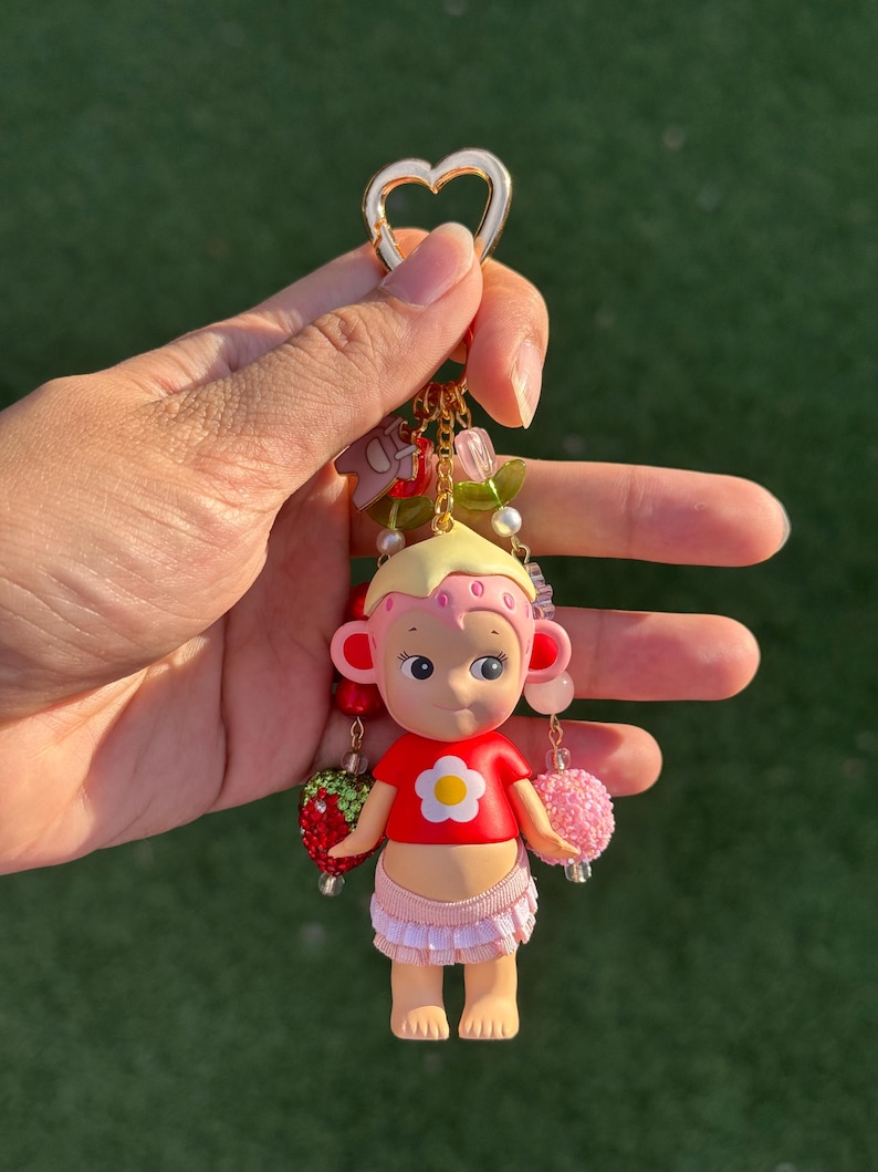 May include: A keychain featuring a small figurine with blonde hair, a pink strawberry-patterned headpiece, and a red shirt with a white flower graphic. The figure wears a light pink ruffled skirt and is accented with dangling beads, a strawberry charm, and a heart-shaped gold clasp.