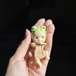 May include: A small figurine of a baby wearing a light green frog hat with pink flower accents. The figure has a pale skin tone, large dark eyes, and a small smile. The text "Sonny Angel" and "DREAMS CHINA" is embossed on the chest area of the figure.