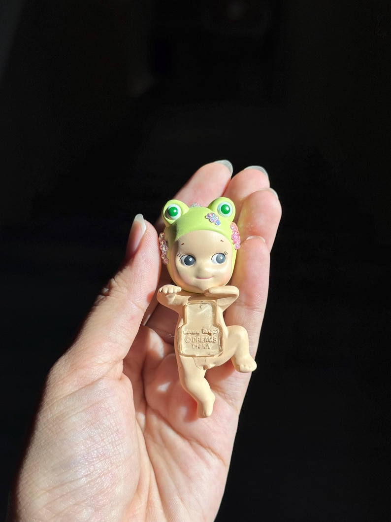 May include: A small figurine of a baby wearing a light green frog hat with pink flower accents. The figure has a pale skin tone, large dark eyes, and a small smile. The text "Sonny Angel" and "DREAMS CHINA" is embossed on the chest area of the figure.