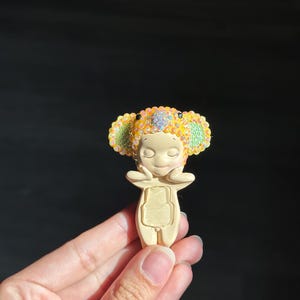 May include: A small, cream-colored figurine of a character wearing a headpiece adorned with sparkling yellow and green rhinestones. The character has closed eyes, a gentle expression, and hands touching its cheeks, with a rectangular cutout on its torso.
