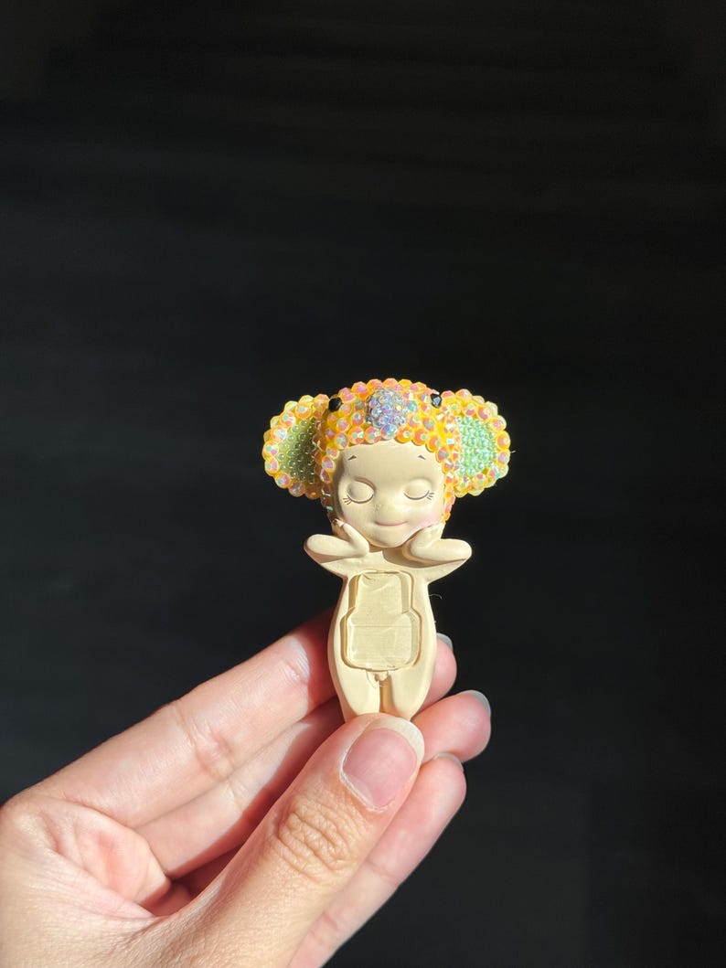 May include: A small, cream-colored figurine of a character wearing a headpiece adorned with sparkling yellow and green rhinestones. The character has closed eyes, a gentle expression, and hands touching its cheeks, with a rectangular cutout on its torso.