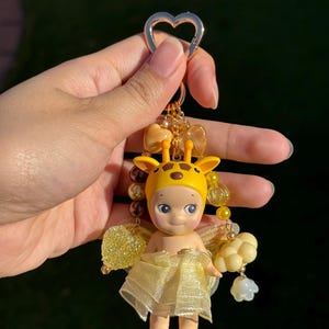 Puede incluir: Un llavero amarillo con tem&aacute;tica de jirafa con una peque&ntilde;a mu&ntilde;eca y un cierre en forma de coraz&oacute;n. La mu&ntilde;eca lleva un sombrero de jirafa amarillo y una falda amarilla transparente. El llavero incluye cuentas y abalorios dorados.