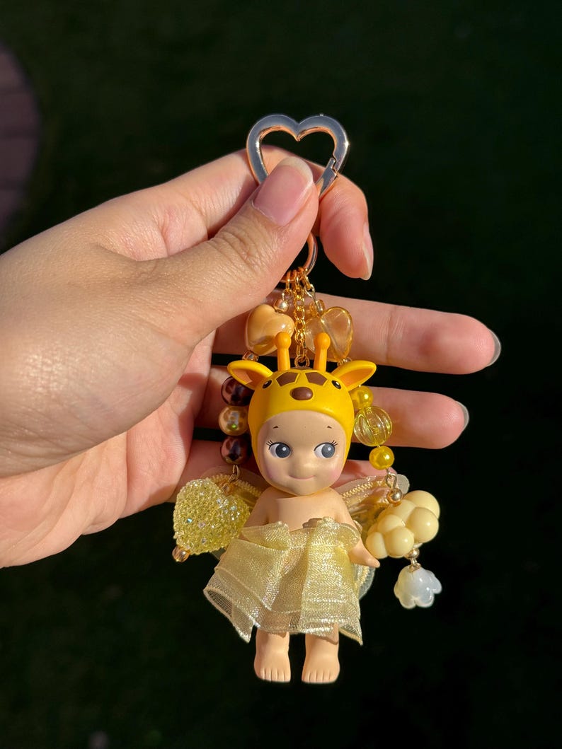 Puede incluir: Un llavero amarillo con tem&aacute;tica de jirafa con una peque&ntilde;a mu&ntilde;eca y un cierre en forma de coraz&oacute;n. La mu&ntilde;eca lleva un sombrero de jirafa amarillo y una falda amarilla transparente. El llavero incluye cuentas y abalorios dorados.