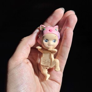 May include: A small, light-skinned figurine wearing a pink cat-themed hood with iridescent accents. The figure has a neutral expression, large eyes, and a pose with arms extended. The text "&copy;DREAMS CHINA" is embossed on the chest area of the figure.