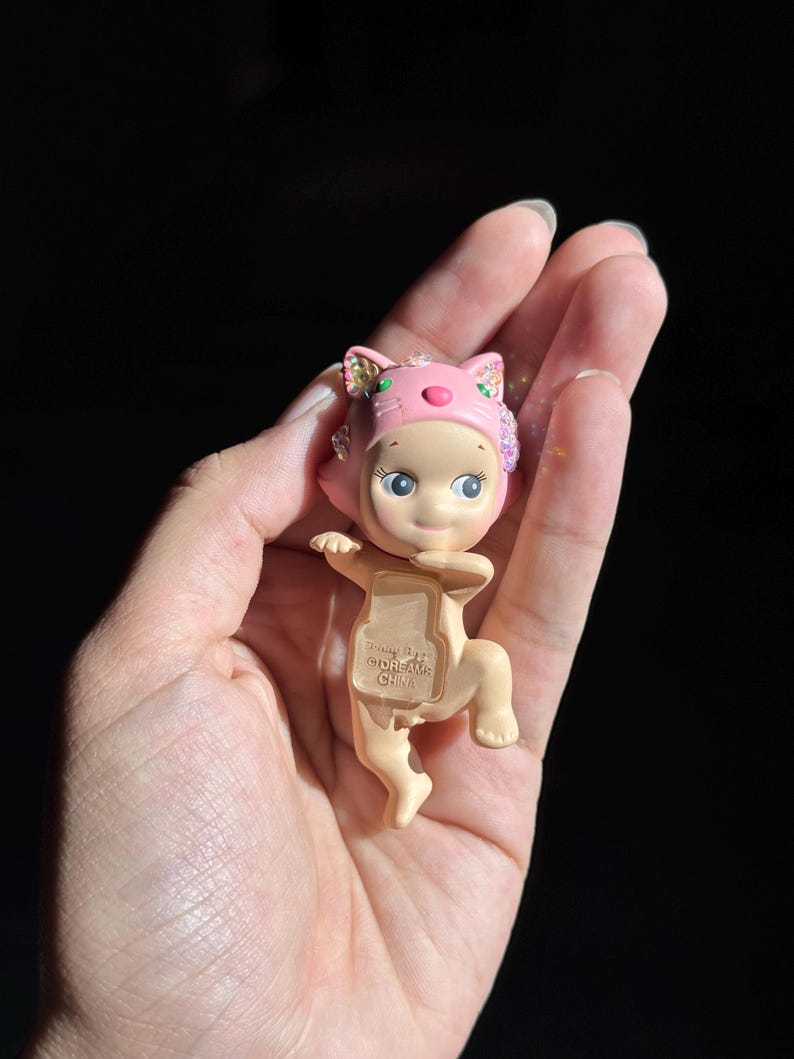 May include: A small, light-skinned figurine wearing a pink cat-themed hood with iridescent accents. The figure has a neutral expression, large eyes, and a pose with arms extended. The text "&copy;DREAMS CHINA" is embossed on the chest area of the figure.