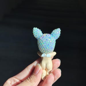 May include: A small figurine featuring a smooth, light-toned body with a pair of white wings on the back. The head is covered in iridescent, light blue, sparkling rhinestones that catch the light, giving it a textured and reflective appearance.