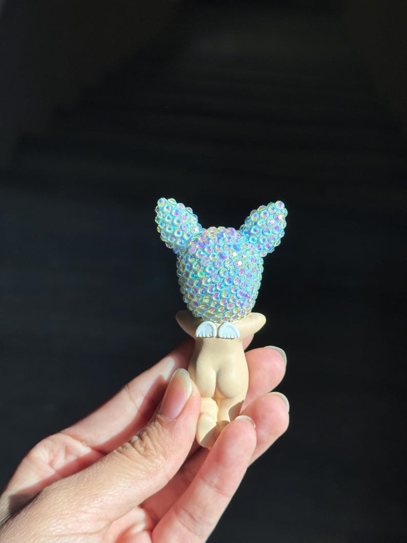 May include: A small figurine featuring a smooth, light-toned body with a pair of white wings on the back. The head is covered in iridescent, light blue, sparkling rhinestones that catch the light, giving it a textured and reflective appearance.